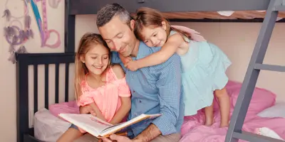 Parent sitting on the bottom bunk of a bed while reading a book. A child sits beside him while another child stand behind him with arms around his neck.