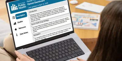 A person views a laptop with the description tab of the Huron Perth opioid dashboard on the screen