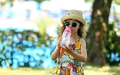 A child wearing a hat and sunglasses and drinking from a water bottle stands outdoors in the shade of a tree.