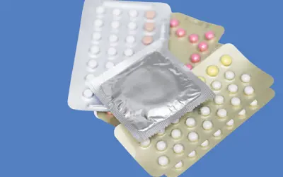 Three packs of birth control pills with a condom in a wrapper sitting on top on blue background